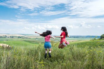 two happy slim smiling young women jumping together on rural field on the blue sky, summer time