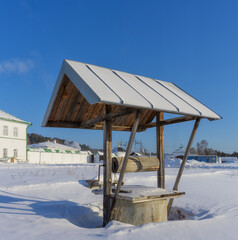 Wooden well for raising water in the snow. The well is traditionally equipped with a drum and a triangular pitched roof. Close-up. Rustic water well against the blue sky. Siberia (Russia), frosty day 