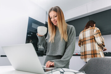 Young woman standing in the kitchen with a mug of coffee, uses a laptop on the background of the girl