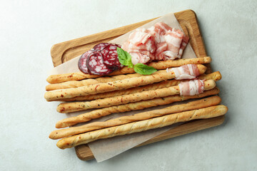 Board with grissini sticks with bacon on white textured background