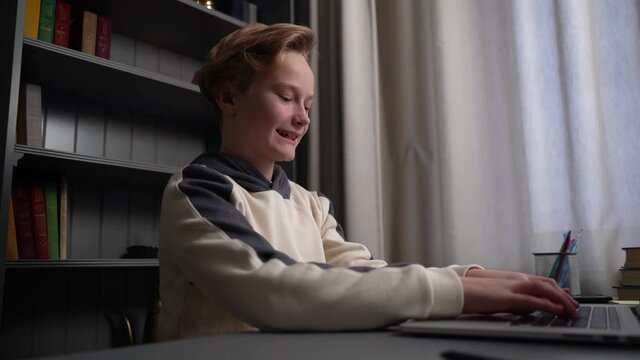 Close-up side view of joyful crazy child boy typing on laptop keyboard sitting at desk near window at home. Laughing kid using computer looking at screen. Tracking shot in slow motion.
