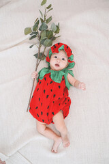 Cute baby girl is wearing a strawberry costume and lying on the bed and holding a branch. top view.