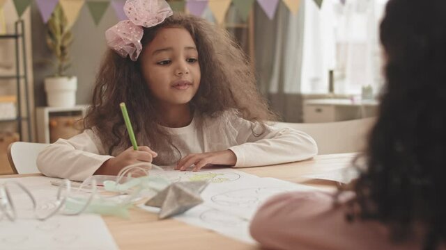 Chest-up of little Mixed-Race girl wearing pink headband with bow, sitting by desk at home, coloring picture with pencil, talking with unrecognizable friend in front of her, then taking toy wand
