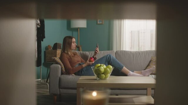 Beautiful girl in blue jeans and brown sweater lies on beige sofa on background of window, biting a red apple and and speaks via video link.
