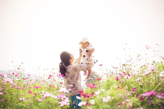 Young Asian Mother With Her Pretty Curly Daughter Are Walking In Spring Garden With Pink Blossom Roses Flowers, Sunset Time. Family Holiday In Summer Day.