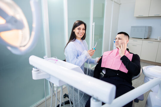 A man sitting in a dental chair, with a worried face holding his hand on his cheek, complains of toothache. Female dentist going to treat a man's tooth