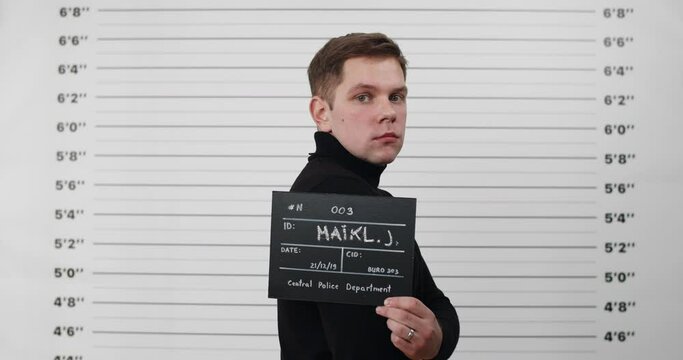 Mugshot of arrested man inblack turtleneck turning head and looking to camera while standing aside . Crop view of young male criminal holding sign in police department.