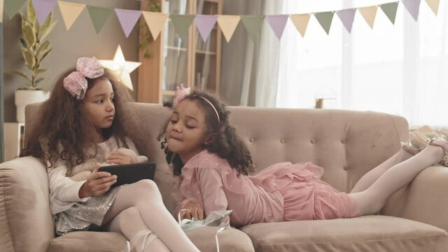 Medium long shot of curly Mixed-Race little girl wearing pretty outfit, sitting on couch in living room, her sister rolling over back of sofa, kids watching smartphone