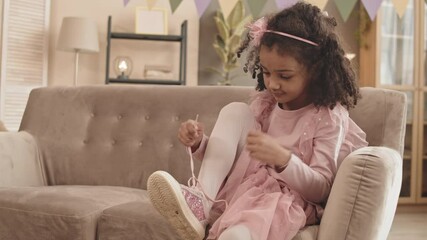Medium long of adorable Mixed-Race little child wearing pink clothing, sitting on sofa in room, trying lacing sparkly shoe