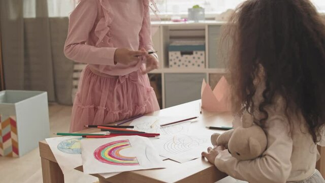 Medium long of two curly little girls sitting at coffee table, doodling over their pictures with pencils