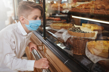 Young boy wearing medical mask looking at desserts on bakery store display, copy space