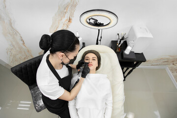 Cosmetologist wearing a mask and gloves applies permanent makeup to the lips of a young girl.