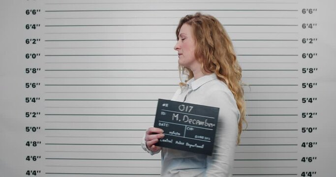 Mugshot of woman in middle 40s with long wavy hair turning to sides while holding sign and posing for photo .Portrait of female beautiful criminal looking to camera in police department.