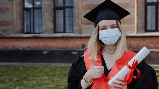 Young graduated girl holding her graduation degree convocation ceremony. Attractive student graduate posing towards the camera in protective mask during coronavirus covid19 background of academy.