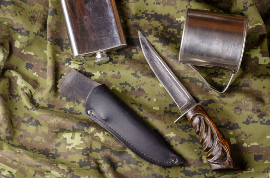 Knife, scabbard, metal flask and mug on camouflage fabric.