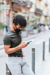 vertical portrait of a latin american young man checking his smart phone in the street. He is wearing casual summer clothes and a black protective face mask.