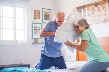 couple of two happy seniors having fun playing together on the bed at home fighting with pillows enjoying - pillows war indoors in the morning. - Powered by Adobe