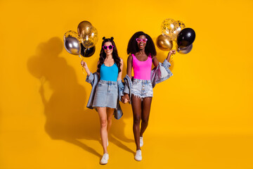 Full length photo of brunette girlfriends walk hold balloons wear singlet denim jacket isolated on yellow color background