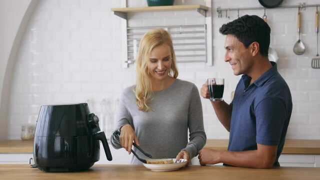 Happy young caucasian husband and wife cooking bread snacks in air fryer for evening snacks with tea at kitchen counter at home