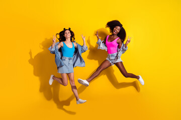 Fototapeta na wymiar Photo of two funny sweet young girlfriends wear denim clothes jumping high showing v-signs isolated yellow color background