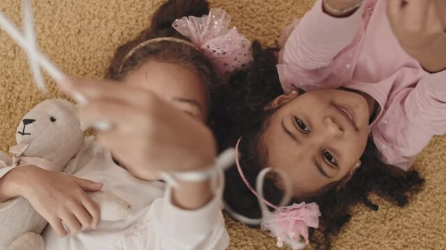 Top view medium POV of two curly Mixed-Race little girls in cute shiny clothes, lying on carpet, waving toy wands, smiling and looking on camera