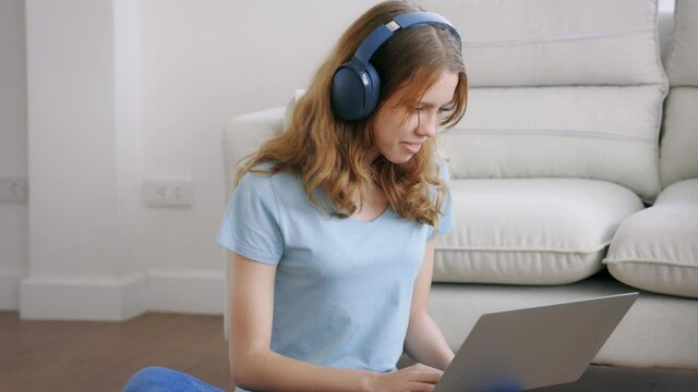 Teenage caucasian girl with bluetooth and wireless headphones using laptop while chatting over smartphone while sitting on carpet in living room