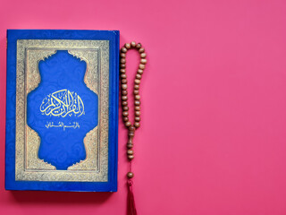 Islamic concept. Selective focus Holy Quran written arabic calligraphy meaning of Al Quran with prayer beads isolated on red background.