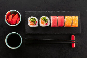 Sushi rolls set, ginger, soy sauce in a bowl and chopsticks from below on a black slate board, top...