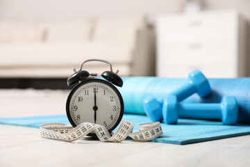 Alarm clock, yoga mat, measuring tape and dumbbells on wooden floor indoors. Morning exercise