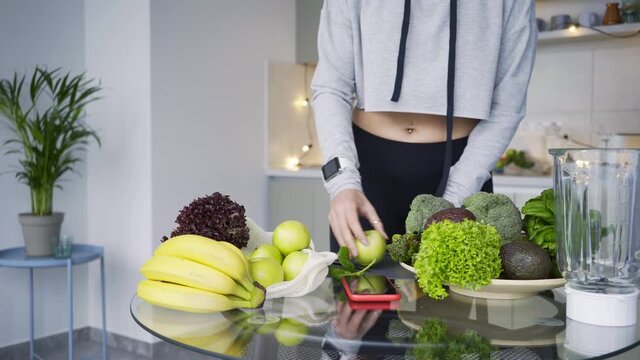 Graceful young sportswoman in stylish tracksuit carries fresh green apples to cook delicious smoothie at glass table in modern kitchen at home