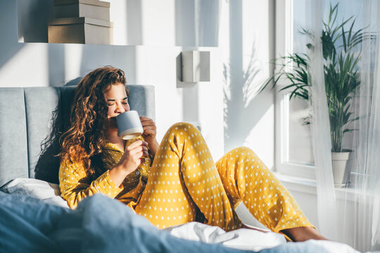 Woman in yellow pajama sitting on bed and drinking coffee at the sunny morning bedroom.