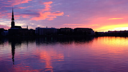 Fototapeta premium Dawn over the Daugava in Riga, the sun illuminates the river, clouds are reflected in the water, the silhouette of Peter's Cathedral is visible