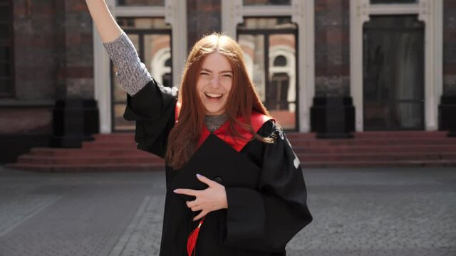 Attractive ginger happy graduate girl student in gown and hat holding diploma laughing rejoices over graduation master's certificate on the background of college university. Education concept.