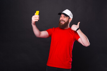 Amazed young bearded hipster man in red t-shirt taking selfie with smartphone over black background and showing thumb up