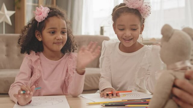 Waist-up of two little adorable Mixed-Race girls sitting at table in living room, waving hands on unrecognizable person holding toy bear in front of them, drawing and smilingWaist-up of two little ado