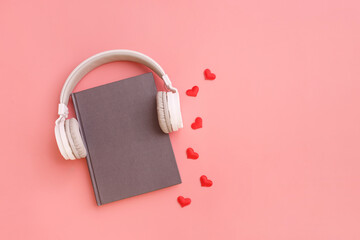 Book with white modern headphones flat lay on pink background with heart. Listen to the audio book concept