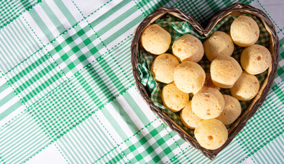 Cheese bread, heart-shaped basket lined with green and white fabric filled with cheese bread on a checkered tablecloth, top view.