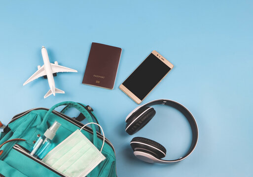 Flat Lay Of Traveling Accessories With Airplane Model And Medical Face Mask And Alcohol Sanitizer Spray In Backpack On Blue Background. Traveling During Covid-19 Epidemic And New Normal.