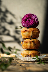 Stack of three homemade cherry and chocolate muffins with ranunculus flower on top on background with shadows.