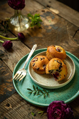 Three homemade cherry and chocolate muffins and ranunculus flowers on ceramic plates on old rustic wooden table. 