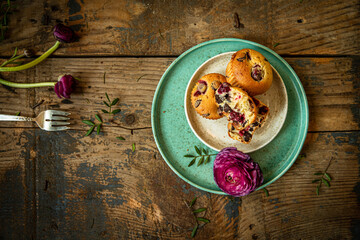 Three homemade cherry and chocolate muffins and ranunculus flowers on ceramic plates on old rustic wooden table. 
