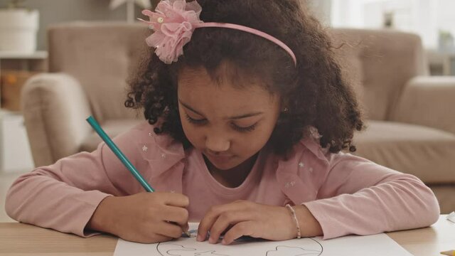 Chest-up POV of curly Mixed-Race five-year old girl wearing pink headband, sitting by desk drawing with pencil, then looking up and smiling on camera