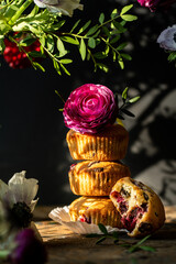 Stack of three homemade cherry and chocolate muffins with ranunculus flower on top and bitten one surrounded with flowers and leaves on black background with shadows.