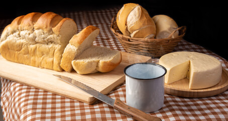 Breakfast table in Brazil with breads, cheese, cup of coffee and accessories on a brown and beige checkered tablecloth, dark background, selective focus.