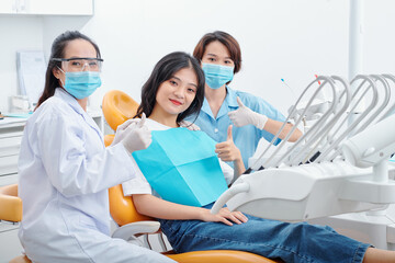 Pretty young Asian woman sitting in dental chair and showing thumbs-up with her dentist and...