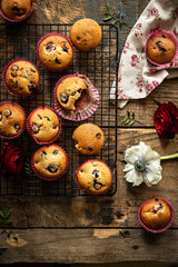 Group of homemade cherry and chocolate muffins on cooling rack and fresh flowers and leaves on rustic wooden table.
