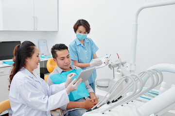Smiling dentist showing tablet computer with teeth x-ray to patient and explaining what to expect from treatment when assistant taking notes in document