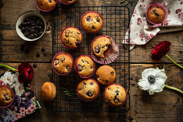 Group of homemade cherry and chocolate muffins on cooling rack and fresh flowers and leaves on rustic wooden table.