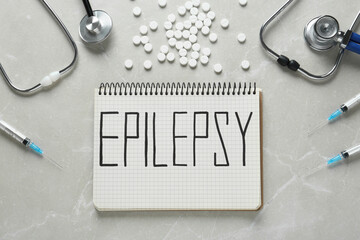 Notebook with word Epilepsy, stethoscopes, pills and syringes on light grey table, flat lay