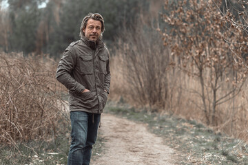 Cheerful blond man in green jacket and jeans on sand path in nature reserve.